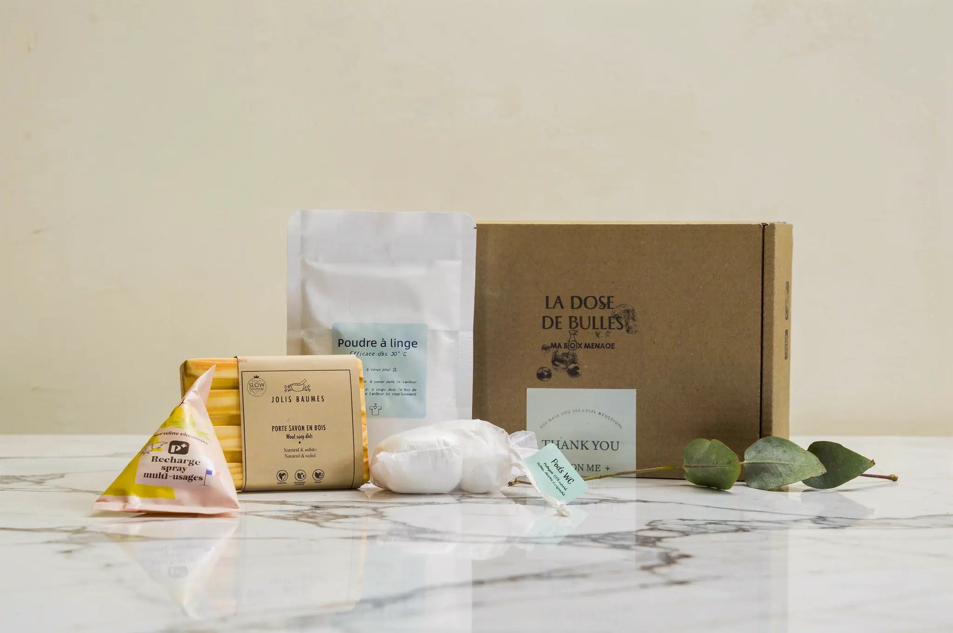Ensemble cadeau de ménage naturel sur marbre: boîte en carton brun "La Dose de Bulles" avec accessoires écologiques (savon bois, sachets, lingette, poudre à linge, emballages compostables) et feuilles vertes.