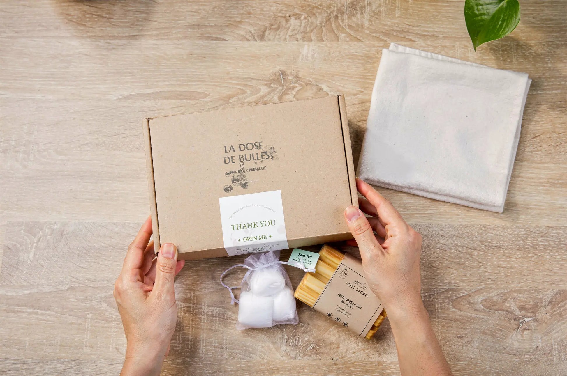 Boîte en carton éco-responsable ouverte montrant des articles de nettoyage naturel: lingettes, sachet de coton, et étiquette “THANK YOU” sur fond bois.