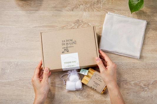 Boîte en carton éco-responsable ouverte montrant des articles de nettoyage naturel: lingettes, sachet de coton, et étiquette “THANK YOU” sur fond bois.