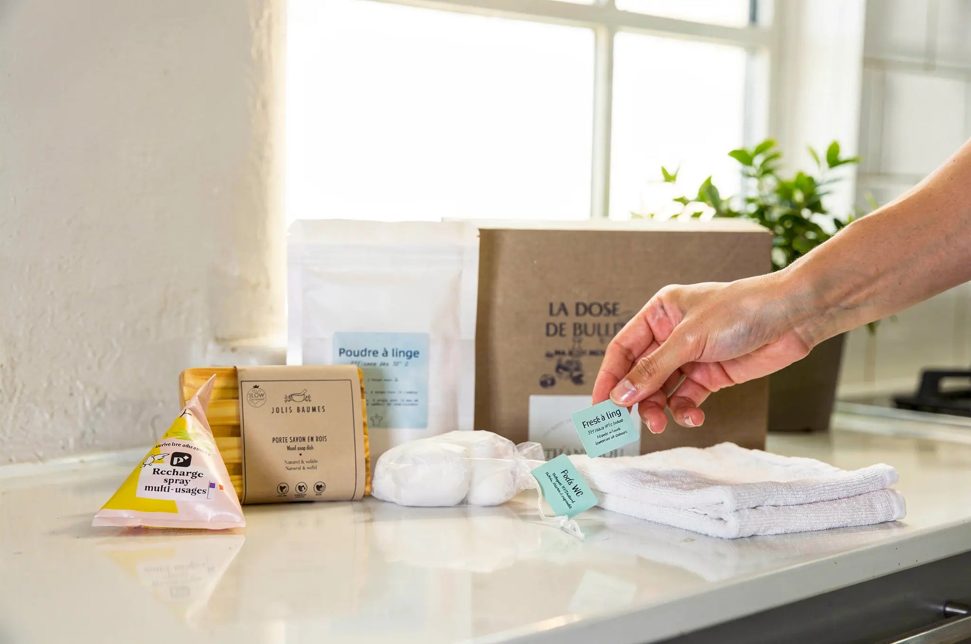 Ensemble de produits ménagers écologiques sur un plan de travail: sachets et emballages en papier, flacons, lingettes et une main choisissant une étiquette bleue, boîte en carton au fond.