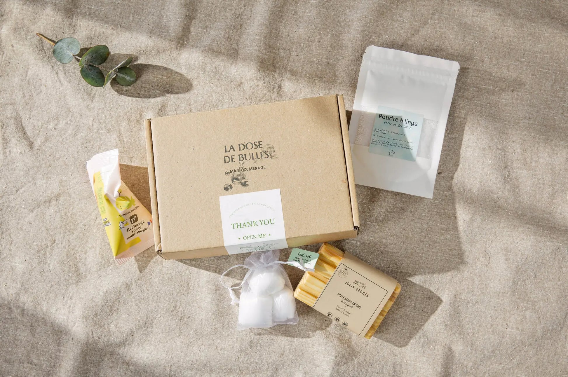 Boîte en carton « Box Essentiel – Nettoyage Naturel Simplifié » entourée de produits écologiques: sachet de lingettes, poudre à linge, savon solide et article en papier beige sur fond beige.