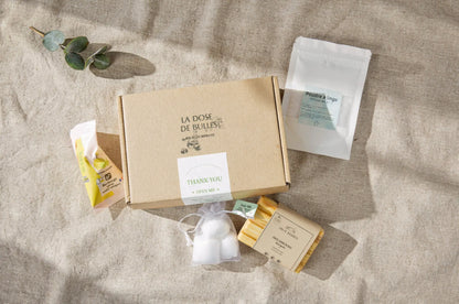 Boîte en carton « Box Essentiel – Nettoyage Naturel Simplifié » entourée de produits écologiques: sachet de lingettes, poudre à linge, savon solide et article en papier beige sur fond beige.
