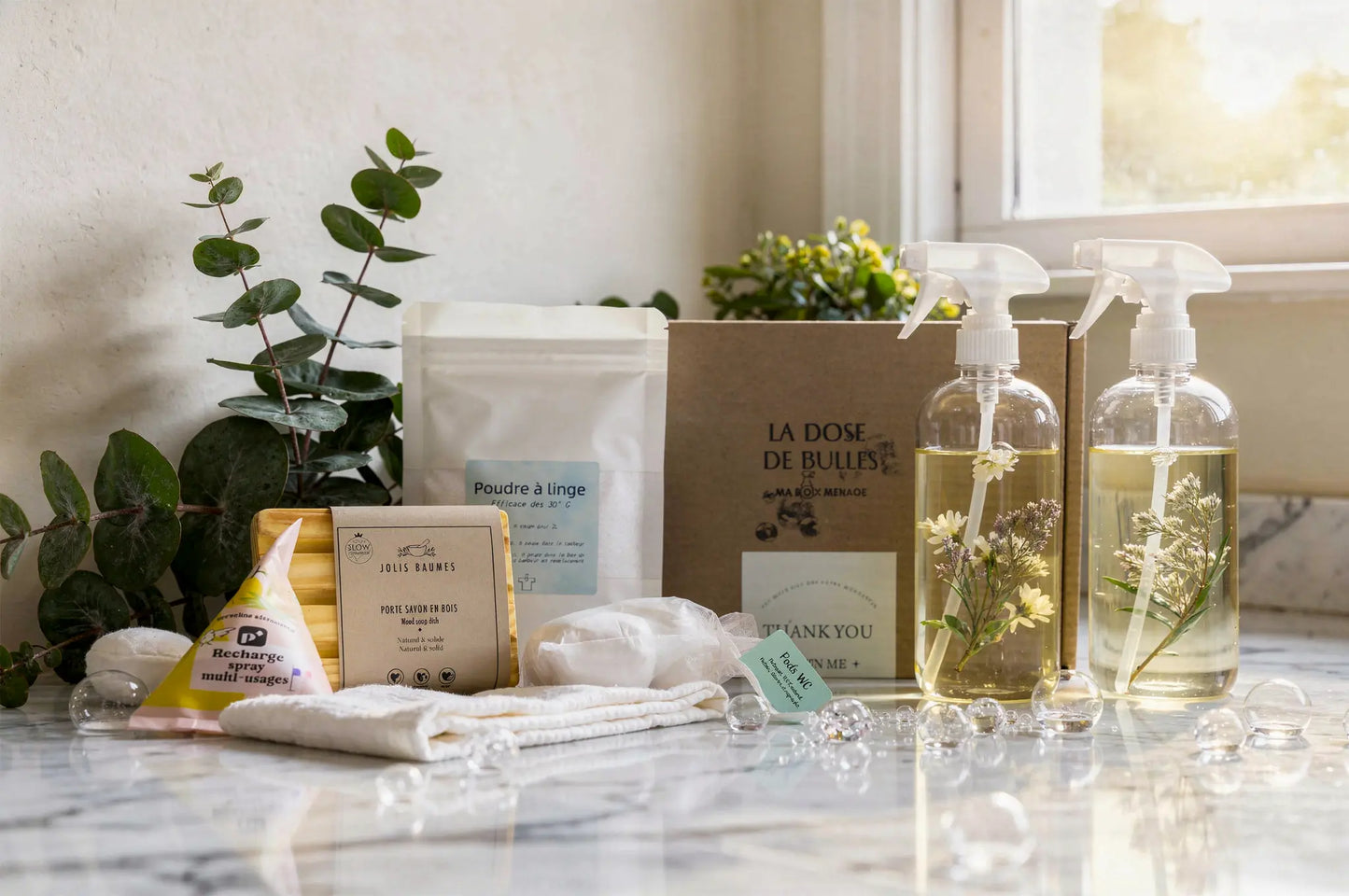 Ensemble de produits ménagers naturels: sprays enzymes dans deux flacons spray avec plantes à l’intérieur, sachets et savon en bois, linge en poudre et accessoires sur une surface marble, ambiance dépouillée et naturelle.