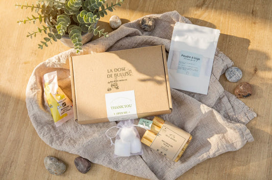 Boîte cadeau éco-nettoyants: carton "La Dose de Bulles" entouré d'accessoires naturels sur lin beige et plantes vertes.
